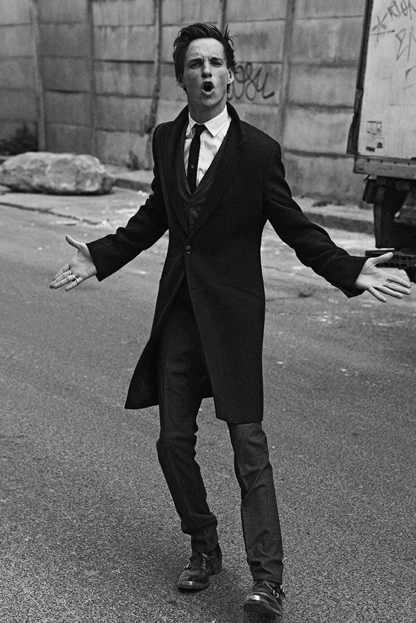 Man in black coat, white shirt, tie, passionately performs with outstretched arms and open mouth.