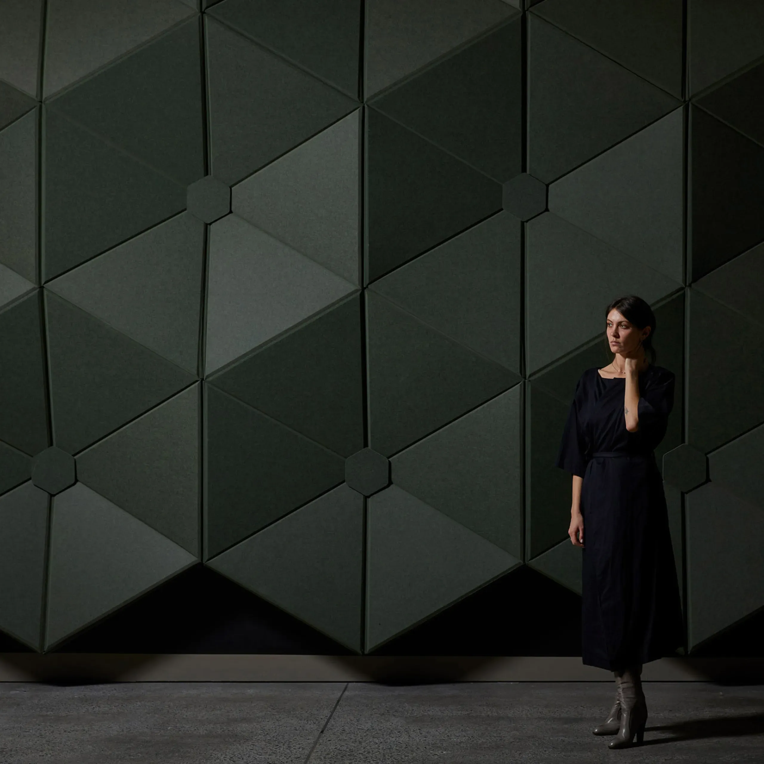 Woman in dark dress stands beside a modern wall with dark green geometric panels.