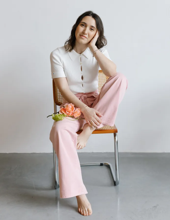 Young woman in white shirt and pink pants sits on a chair, holding orange flowers.