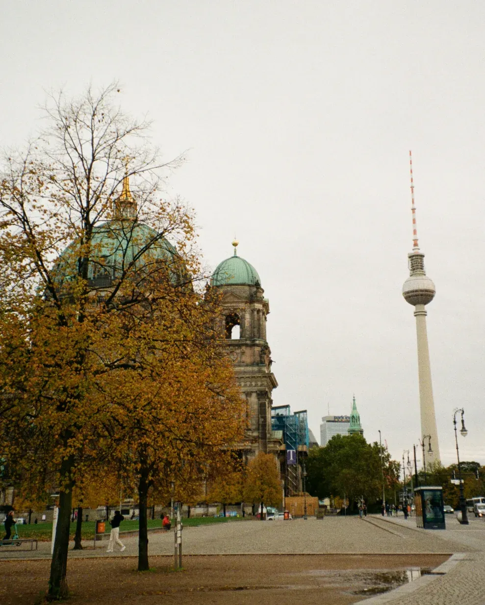 Ornate building with green dome, autumn trees, and the Berlin TV Tower under a grey sky.