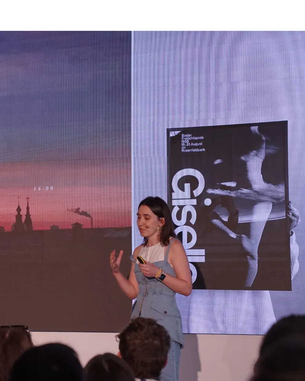 Woman speaking on stage with animated hands, screens show cityscape and 'Giselle' poster.