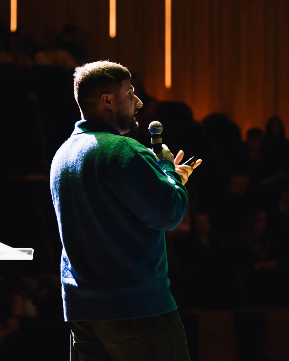 Man in a green sweater speaks passionately into a microphone on a dark stage.