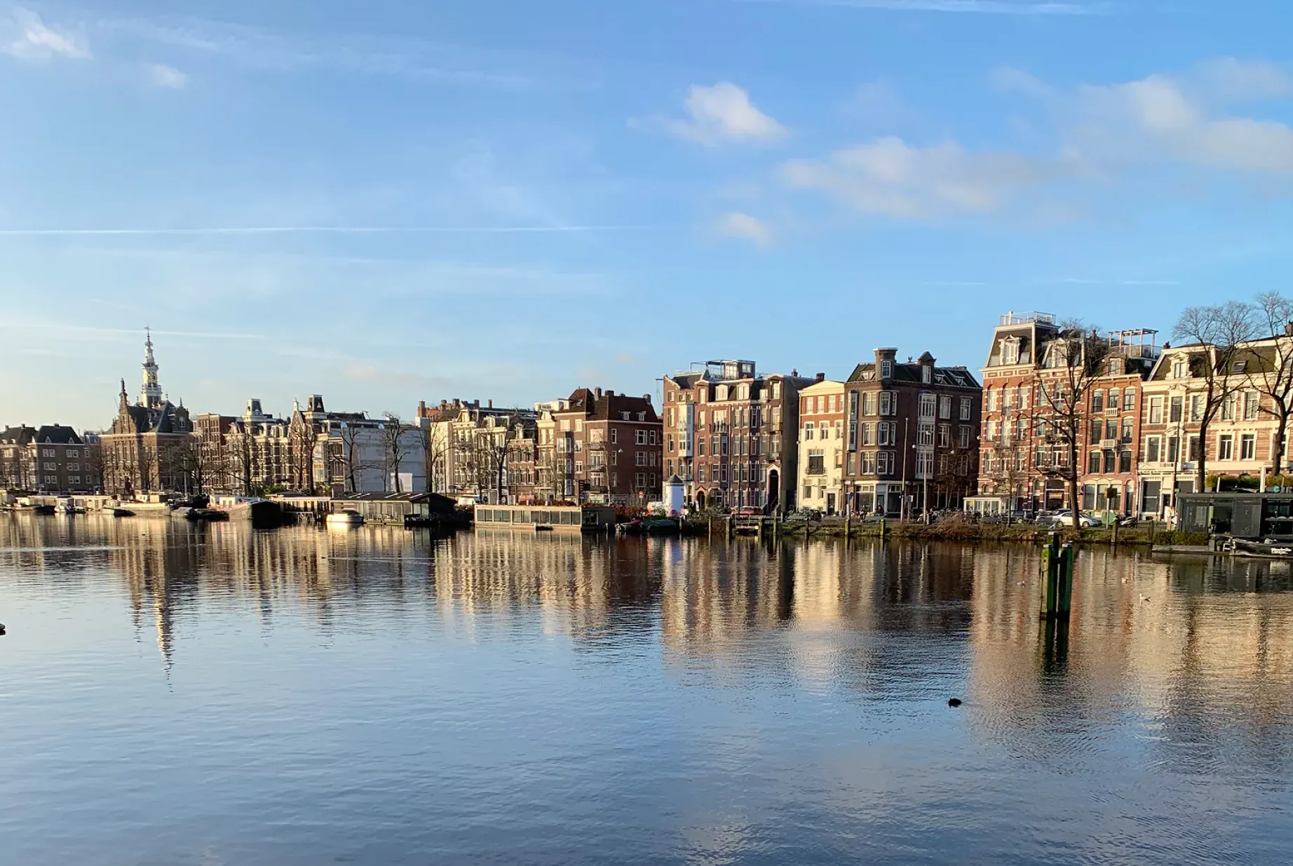 City skyline with historic buildings reflected in calm water under a bright blue sky.