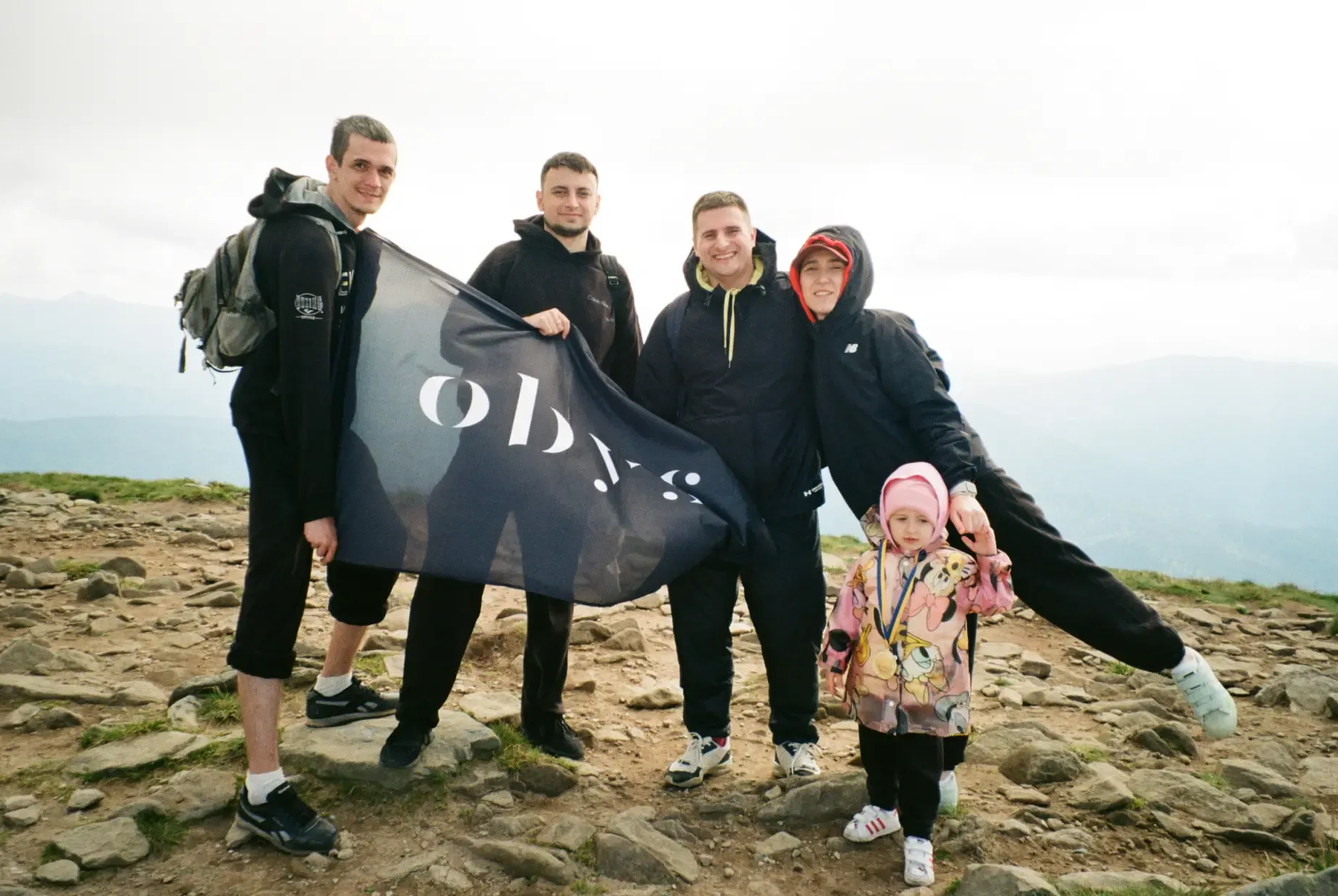 Five people, including a small child, proudly hold a flag on a rocky mountain summit.