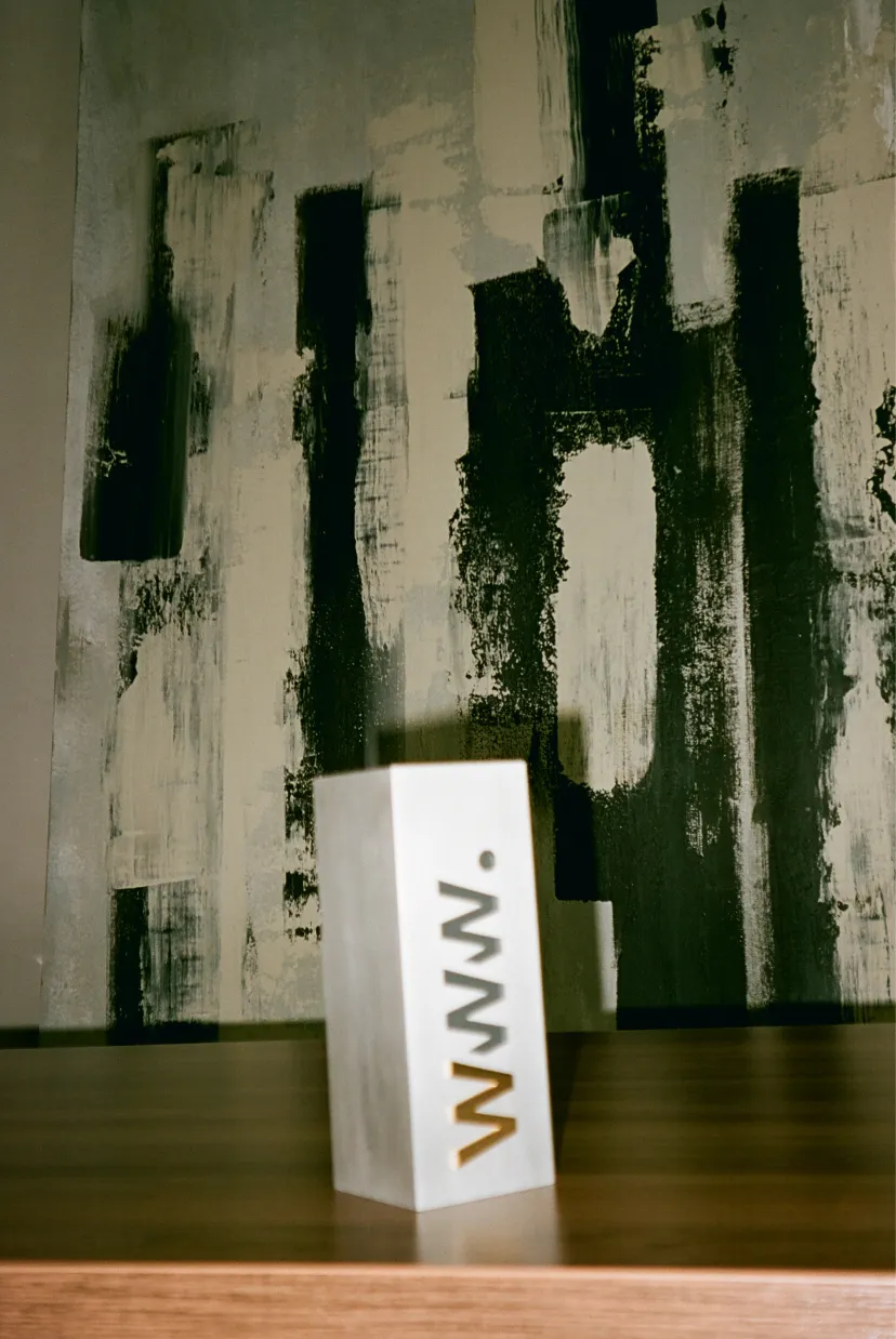 A white block with 'WWW.' printed vertically on its side, against an abstract wall.