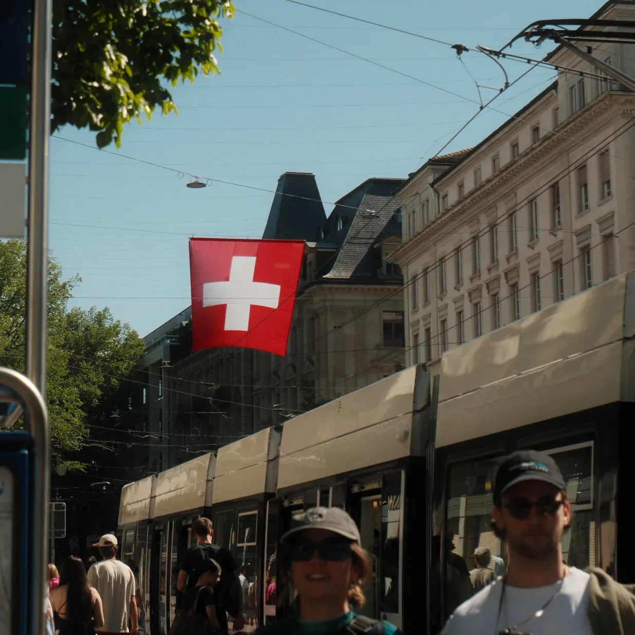 A Swiss flag hangs above a street with a tram and pedestrians in a European city.