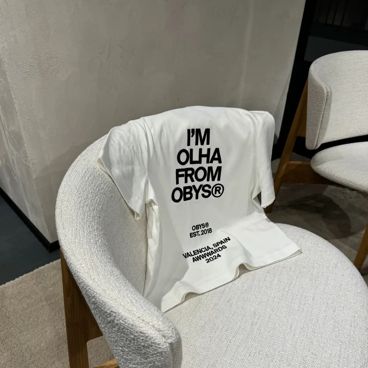 White t-shirt with "I'M OLHA FROM OBYS®" text draped over a textured white chair.